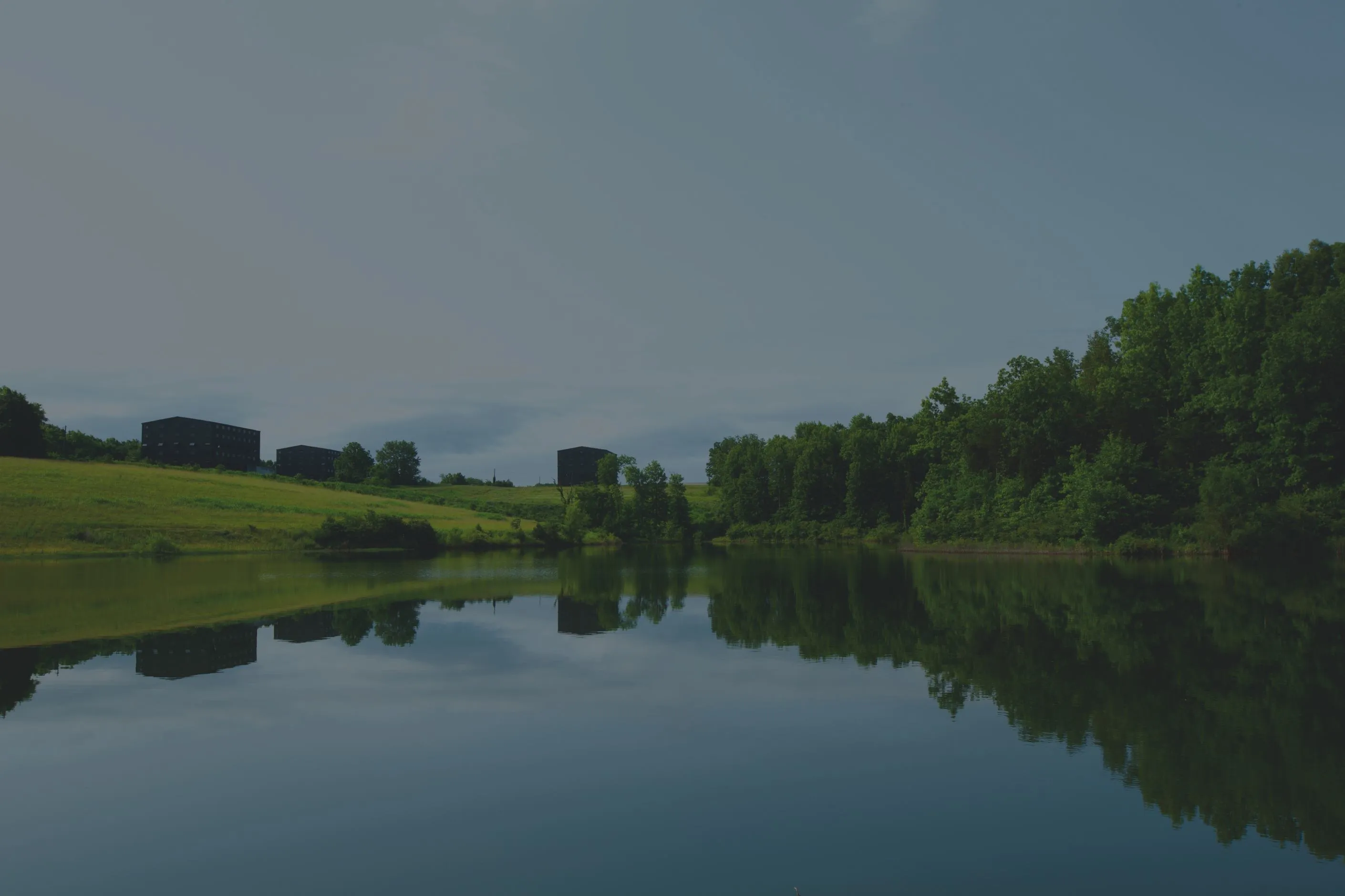 A wide expanse of water showing the reflection of trees and the sky. Beyond the banks of the lake are some large distillery buildings. 